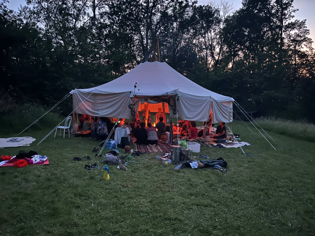 Ein großes weißes Zelt auf einer Wiese in der Abenddämmerung, innen sitzen Menschen im warmen Licht eines Feuers